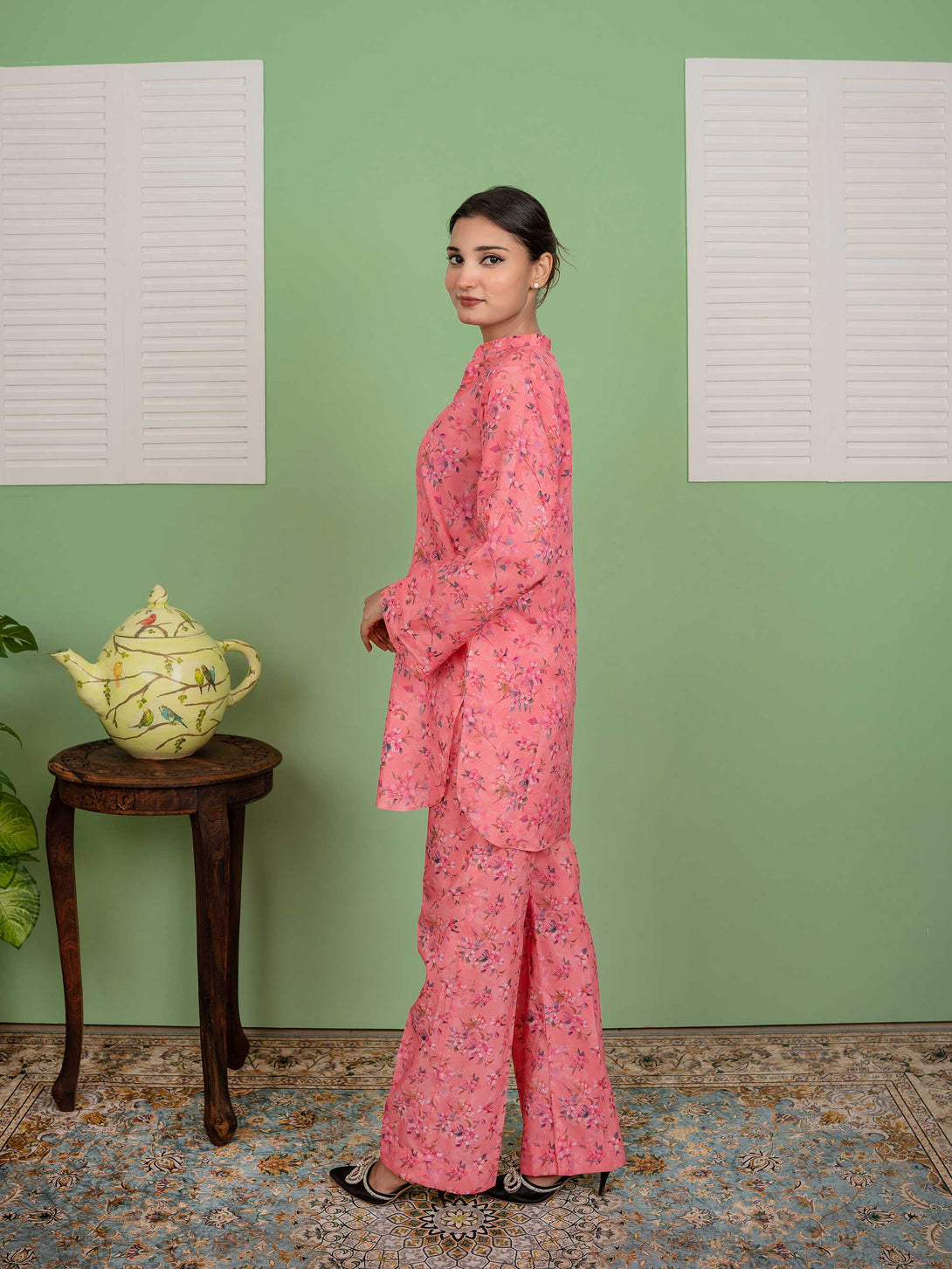 Woman in a pink floral outfit standing in a room with a green wall and wooden stool.